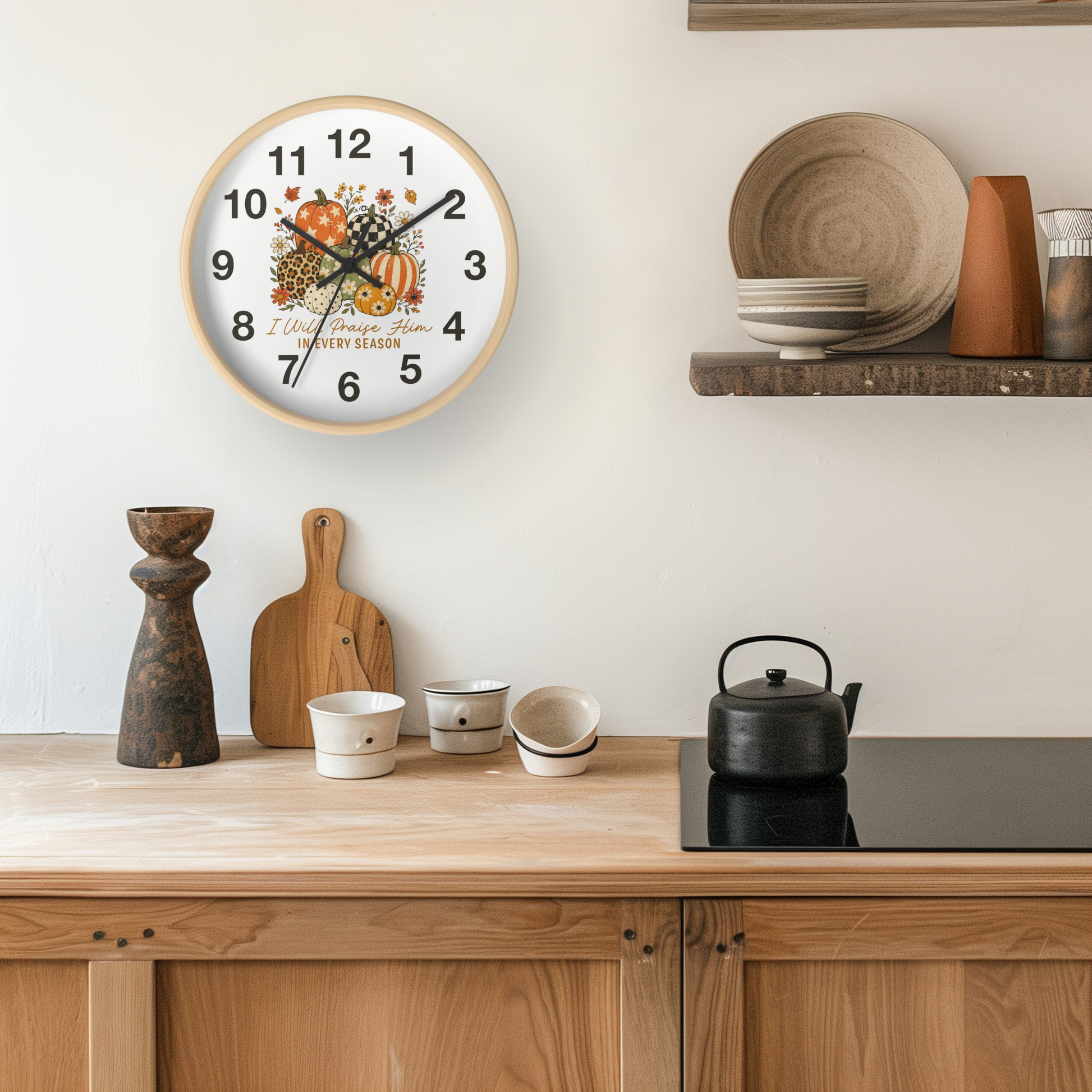 A round wall clock with a fall harvest design hangs on a kitchen wall above a wooden countertop with ceramic cups, a cutting board, and a black kettle.