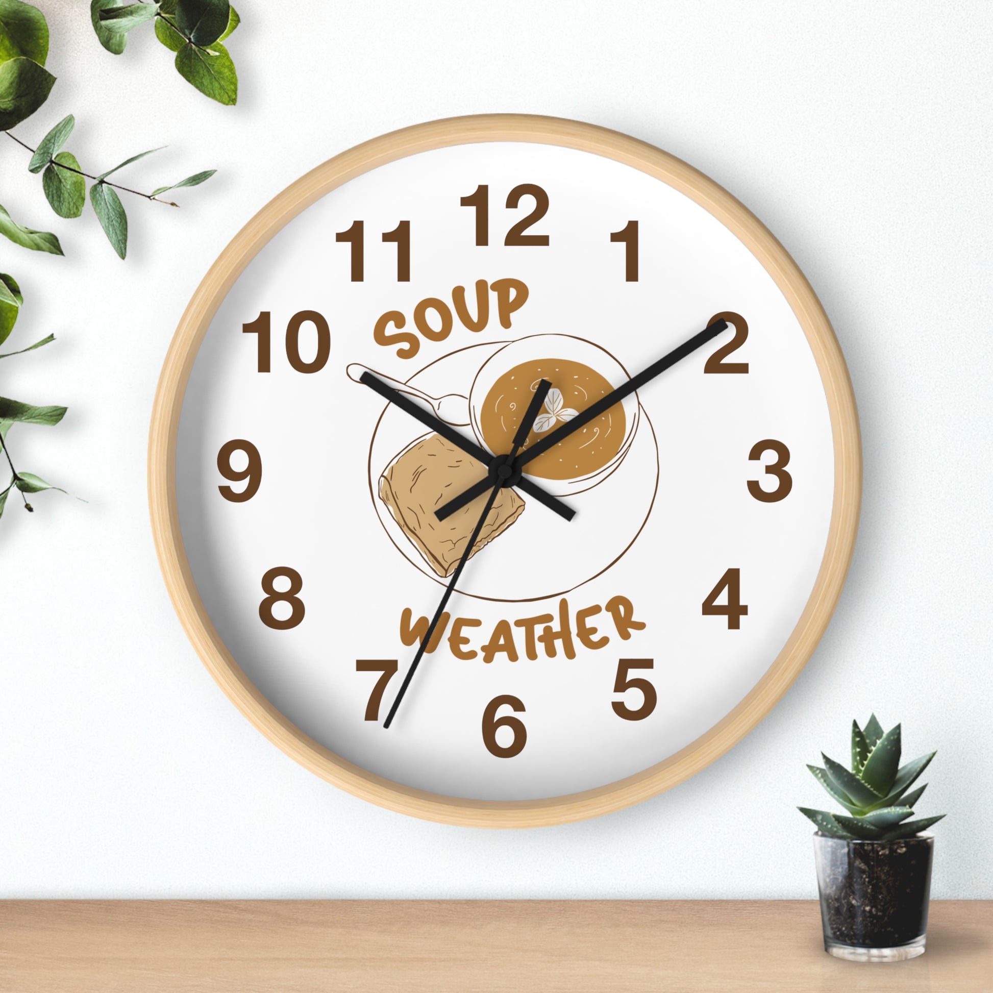 A round wall clock with a light wood frame and a white face, featuring a 'Soup Weather' graphic with a bowl of soup and a cloud. The clock is styled on a light wall above a wooden surface with a small succulent and green leaves.