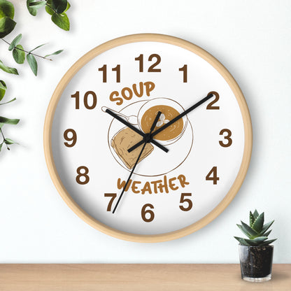 A round wall clock with a light wood frame and a white face, featuring a 'Soup Weather' graphic with a bowl of soup and a cloud. The clock is styled on a light wall above a wooden surface with a small succulent and green leaves.