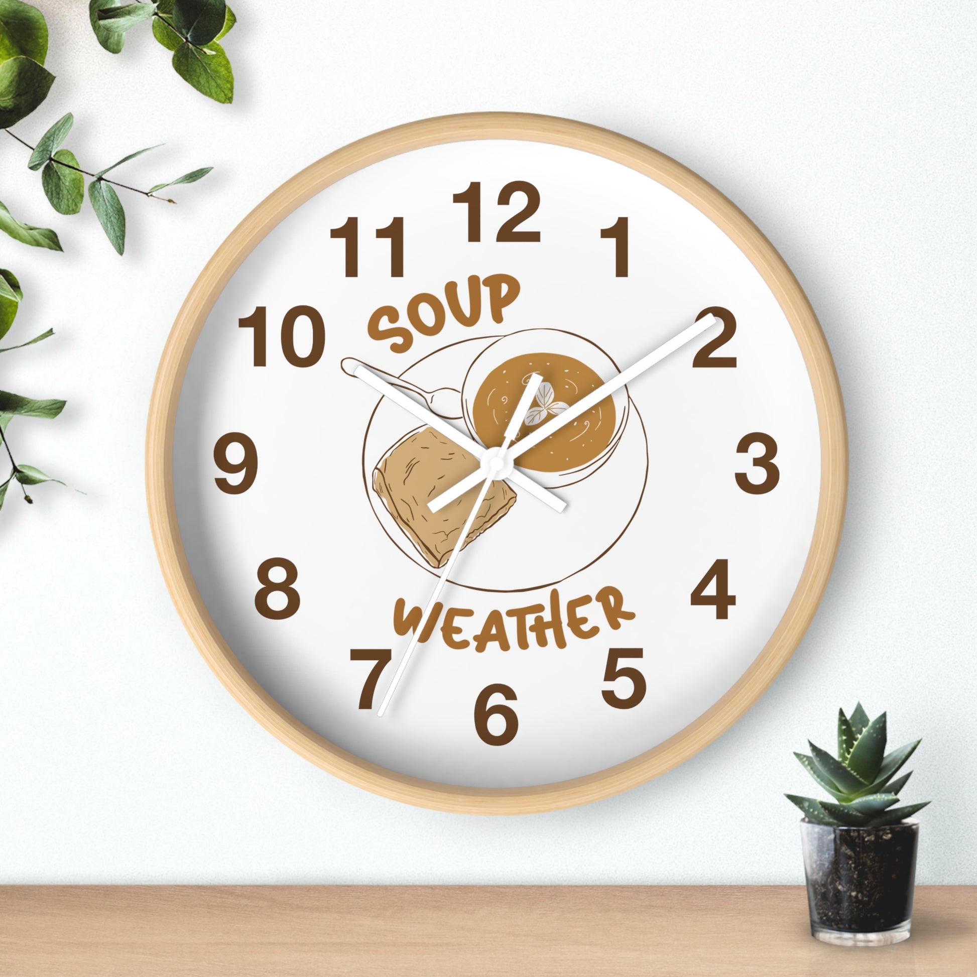 A round wall clock with a light wood frame and a white face, featuring a peanut butter and jelly sandwich illustration with the words 'SOUP' and 'WEATHER' in brown. The clock is shown hanging on a white wall above a wooden surface, with decorative green leaves and a small potted succulent nearby.