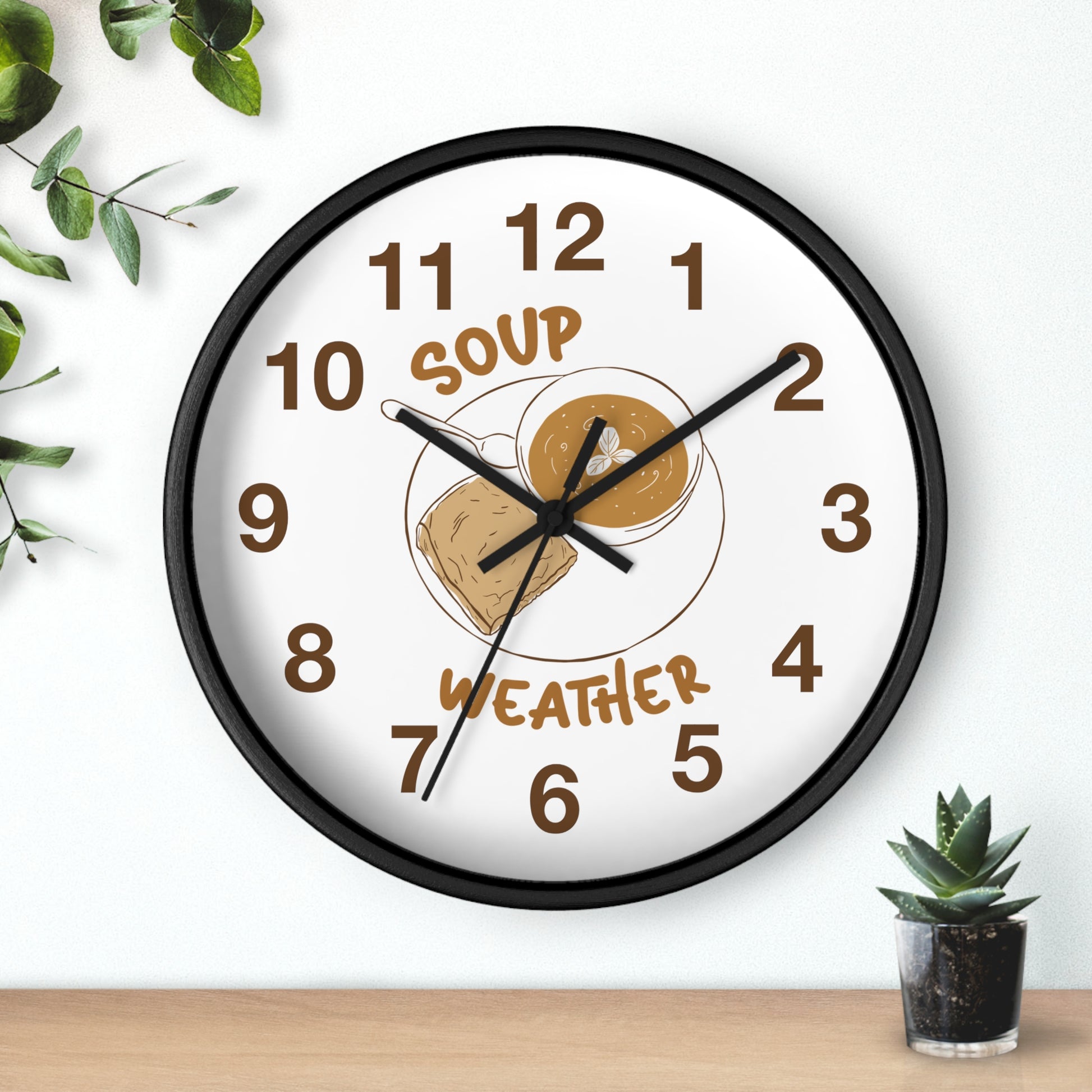 A round wall clock with a black frame and a white face, featuring a 'Soup Weather' graphic with a bowl of soup and a cloud. The clock is styled on a light wall above a wooden surface with small green plants.