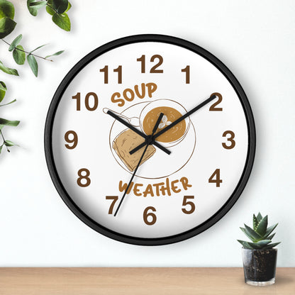 A round wall clock with a black frame and a white face, featuring a 'Soup Weather' graphic with a bowl of soup and a cloud. The clock is styled on a light wall above a wooden surface with small green plants.