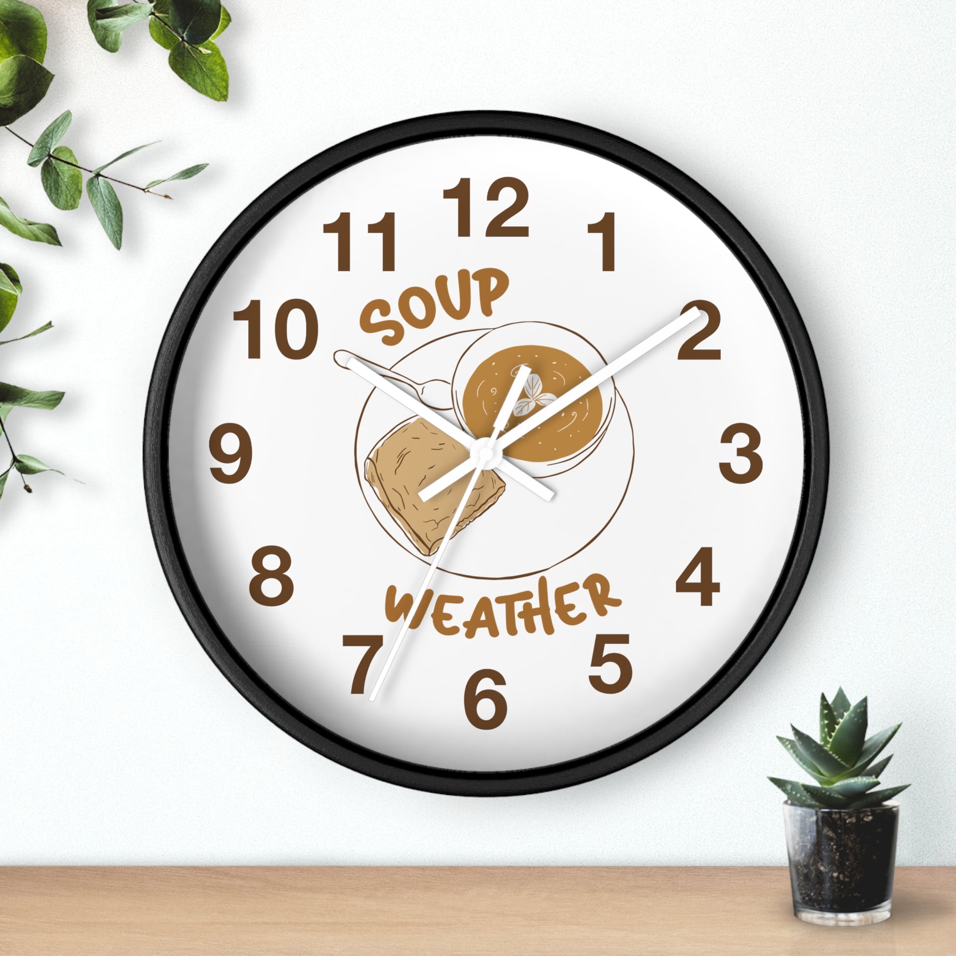 A round wall clock with a black frame and a white face, featuring a peanut butter and jelly sandwich illustration in the center with the words 'SOUP' and 'WEATHER' in brown. The clock is mounted on a white wall above a light wood surface, with green leaves and a small potted succulent nearby.
