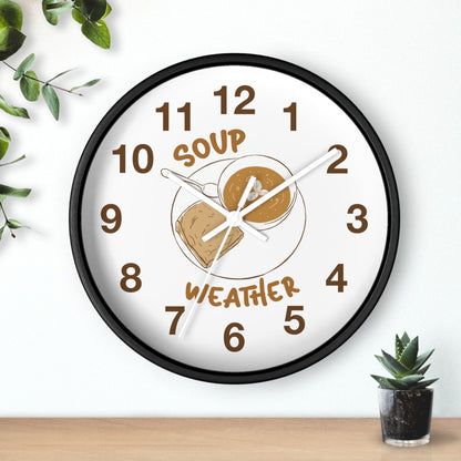 A round wall clock with a black frame and a white face, featuring a peanut butter and jelly sandwich illustration in the center with the words 'SOUP' and 'WEATHER' in brown. The clock is mounted on a white wall above a light wood surface, with green leaves and a small potted succulent nearby.