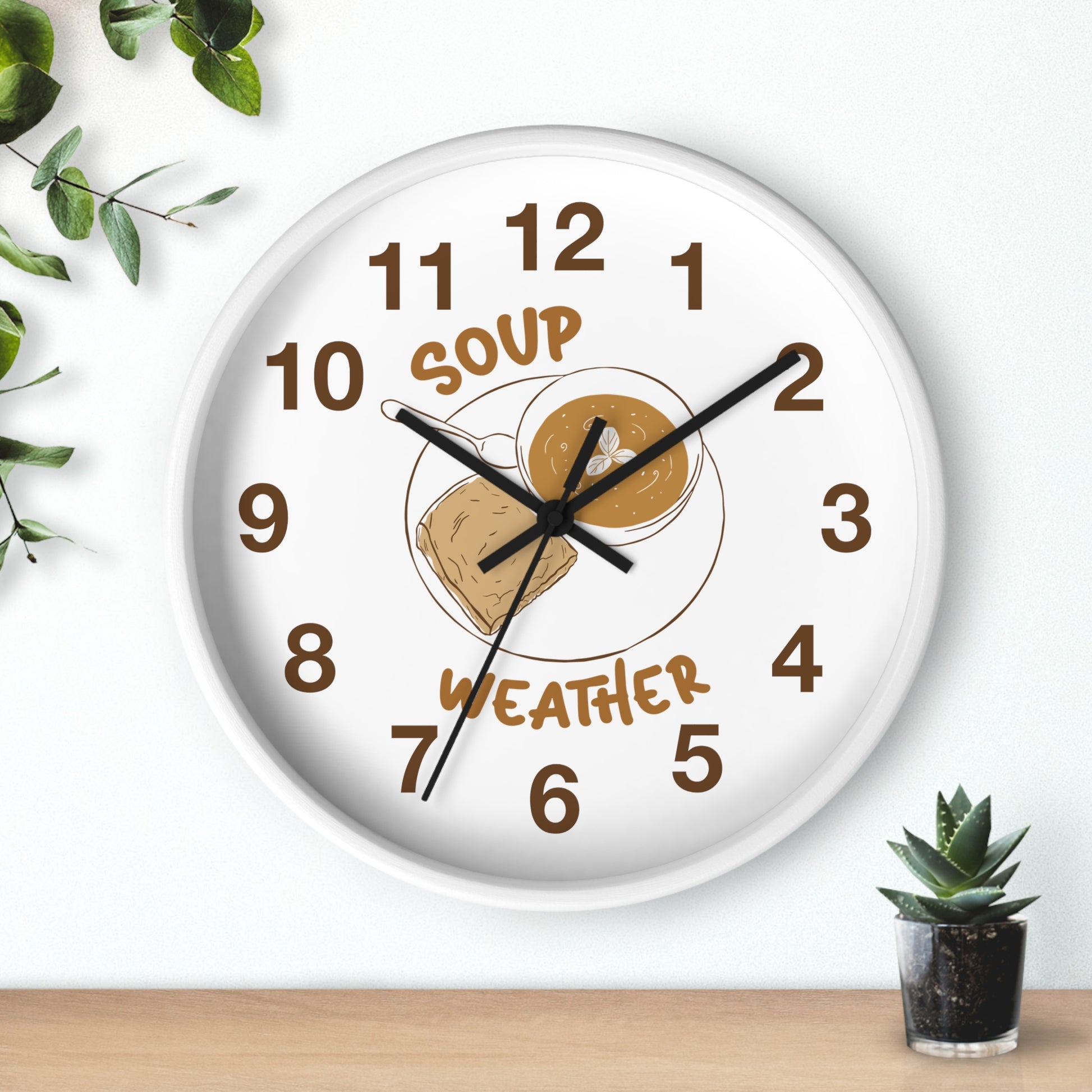 A round white wall clock with a humorous 'Soup Weather' face, featuring brown numerals and a graphic of a bowl of soup with a cloud. The clock is mounted on a white wall above a wooden surface with a small succulent plant and green leaves in the corners.