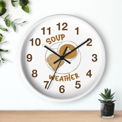 A round white wall clock with a humorous 'Soup Weather' face, featuring brown numerals and a graphic of a bowl of soup with a cloud. The clock is mounted on a white wall above a wooden surface with a small succulent plant and green leaves in the corners.