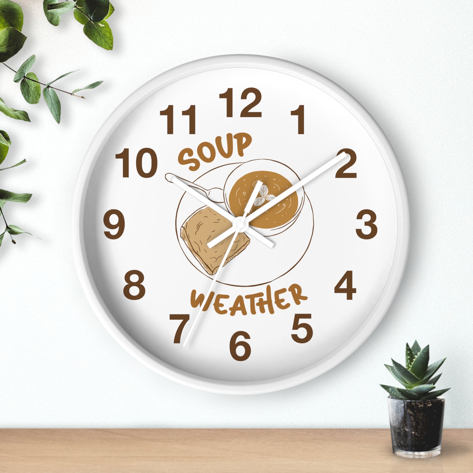 A round white wall clock with a humorous 'Soup Weather' face design, featuring brown numerals and an illustration of a bowl of soup and a slice of toast. The clock is mounted on a white wall above a light wood surface, styled with a small potted succulent and green leaves in the corners.