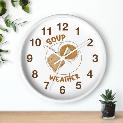 A round white wall clock with a humorous 'Soup Weather' face design, featuring brown numerals and an illustration of a bowl of soup and a slice of toast. The clock is mounted on a white wall above a light wood surface, styled with a small potted succulent and green leaves in the corners.