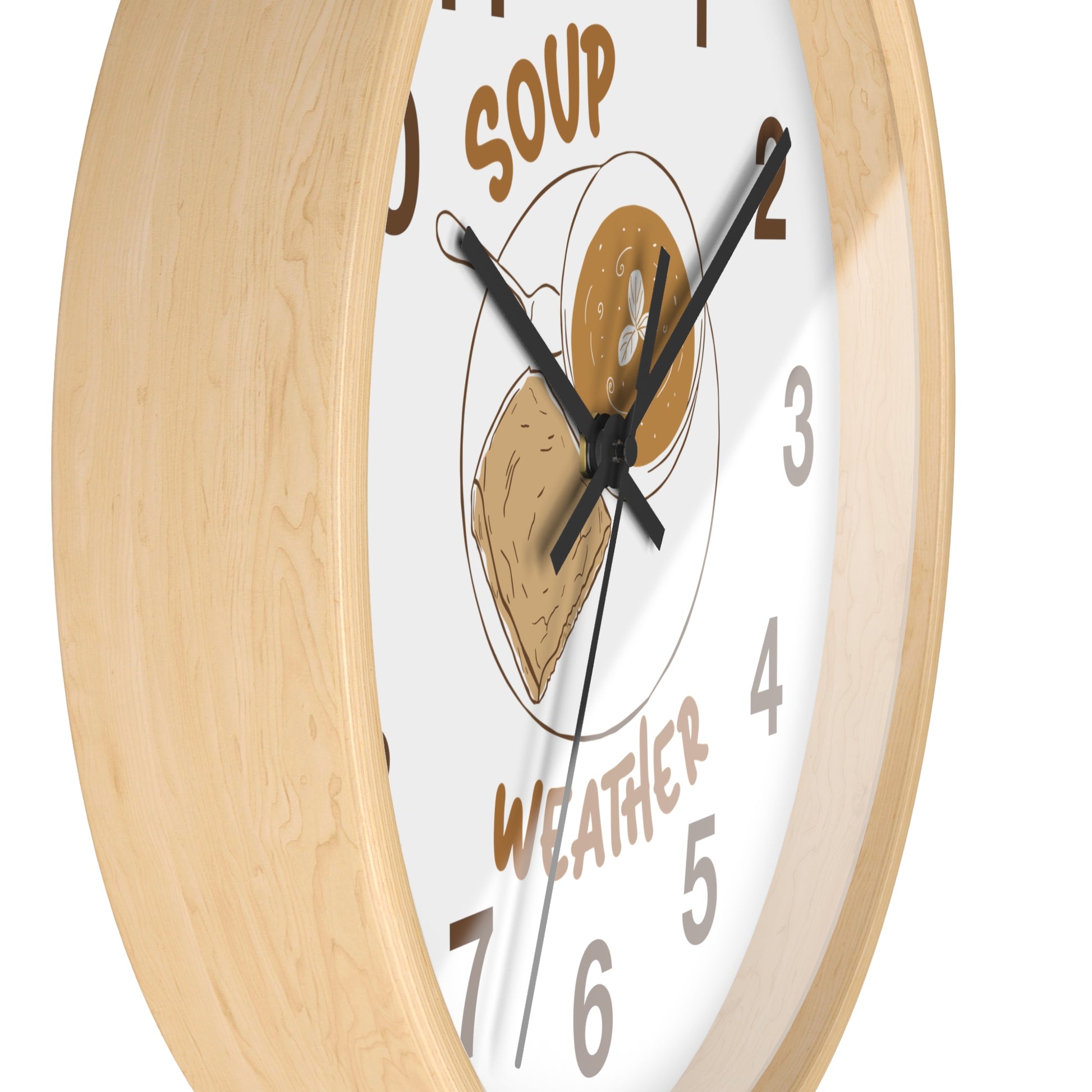 A close-up of a round wall clock with a light natural wood frame and a white face. The clock face features a whimsical illustration of a soup bowl with a slice of toast and a piece of bread, with the words 'SOUP' and 'WEATHER' in brown text, and black numerals.