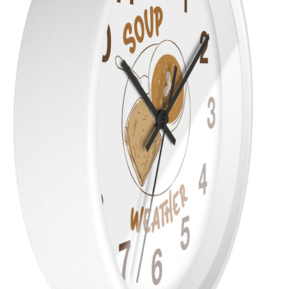A white round wall clock with a whimsical food-themed face. The center features an illustration of a bowl of soup and a slice of toast, with the words 'SOUP' and 'WEATHER' in brown lettering, and gray numerals marking the hours.
