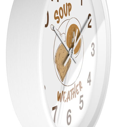 A white round wall clock with a whimsical 'Soup Weather' design. The clock face features brown text reading 'SOUP' at the top and 'WEATHER' at the bottom, with a central illustration of a bowl containing a slice of toast and a spoonful of soup.