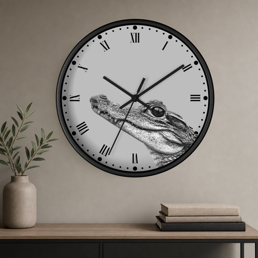 A round wall clock with a black frame and a white face featuring a detailed black-and-white crocodile illustration. The clock is mounted on a beige wall above a wooden console table with a vase and books.