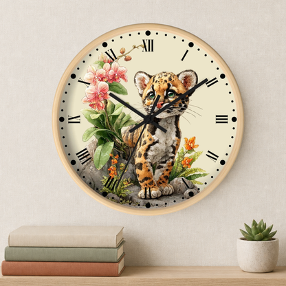 A round wall clock with a light wood frame and a cream dial, featuring an illustration of a spotted big cat surrounded by pink flowers and green leaves. The clock is mounted on a light beige wall above a wooden shelf with stacked books and a small potted succulent.