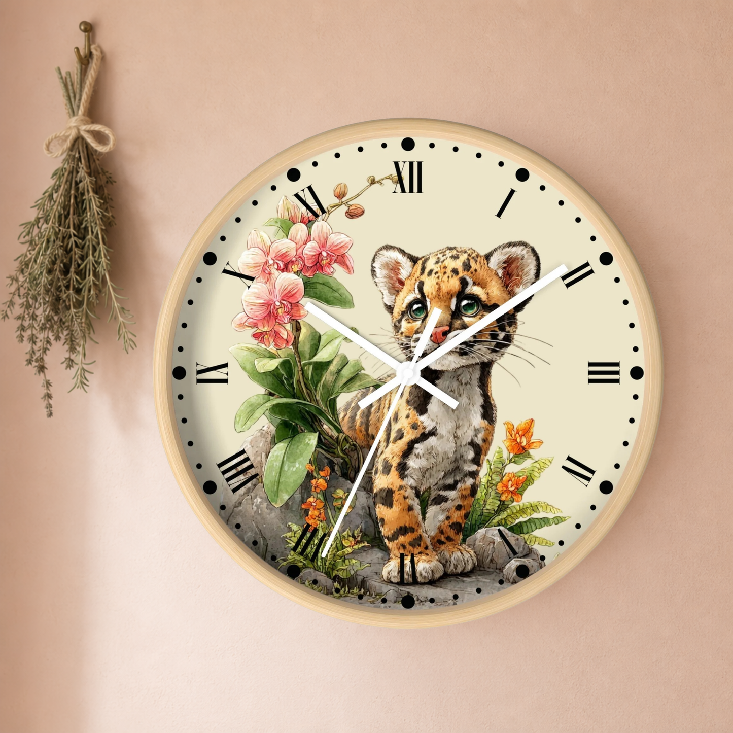 A round wall clock with a natural wood frame and a cream-colored face, featuring an illustration of a spotted Ocelot cat sitting among pink flowers and green leaves. The clock is mounted on a warm beige wall with a dried herb sprig hanging to the left.