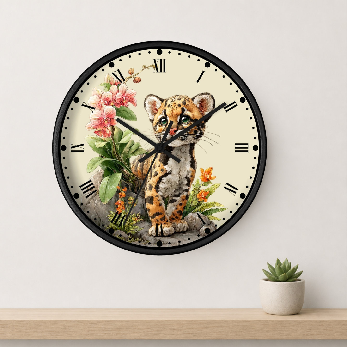 A round wall clock with a black frame and a cream-colored face, featuring an illustration of a spotted kitten surrounded by pink flowers and green leaves. The clock is mounted on a light gray wall above a wooden shelf with a small potted succulent.