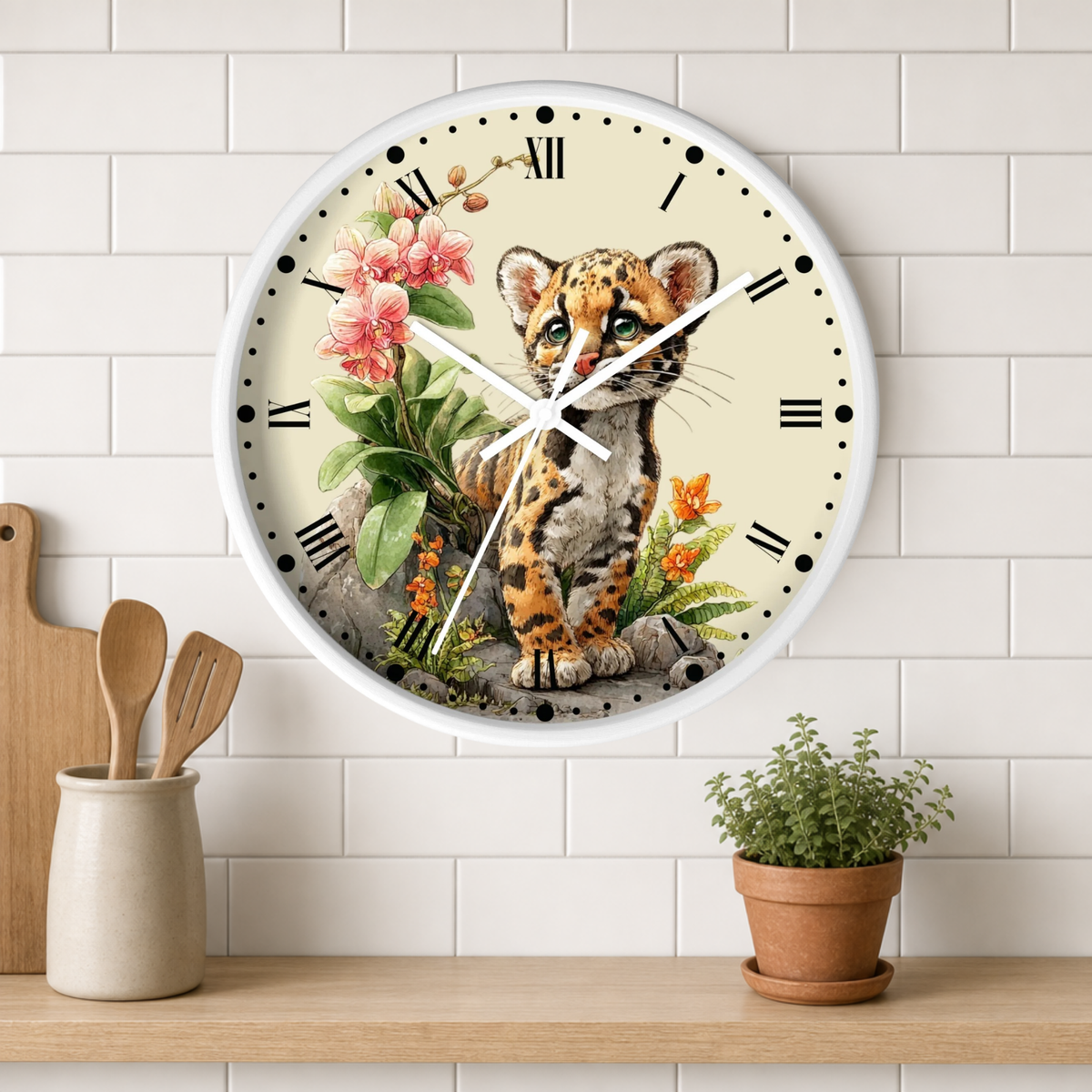 A round wall clock with a white frame and a detailed illustration of a spotted big cat sitting among pink flowers and green leaves on the face. The clock is mounted on a white subway-tile wall above a wooden shelf with a small potted plant and wooden utensils.