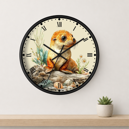 A round wall clock with a black frame and a cream-colored face featuring a detailed illustration of a seal pup sitting on a log with small plants. The clock is mounted on a light gray wall above a wooden shelf with a small potted succulent.