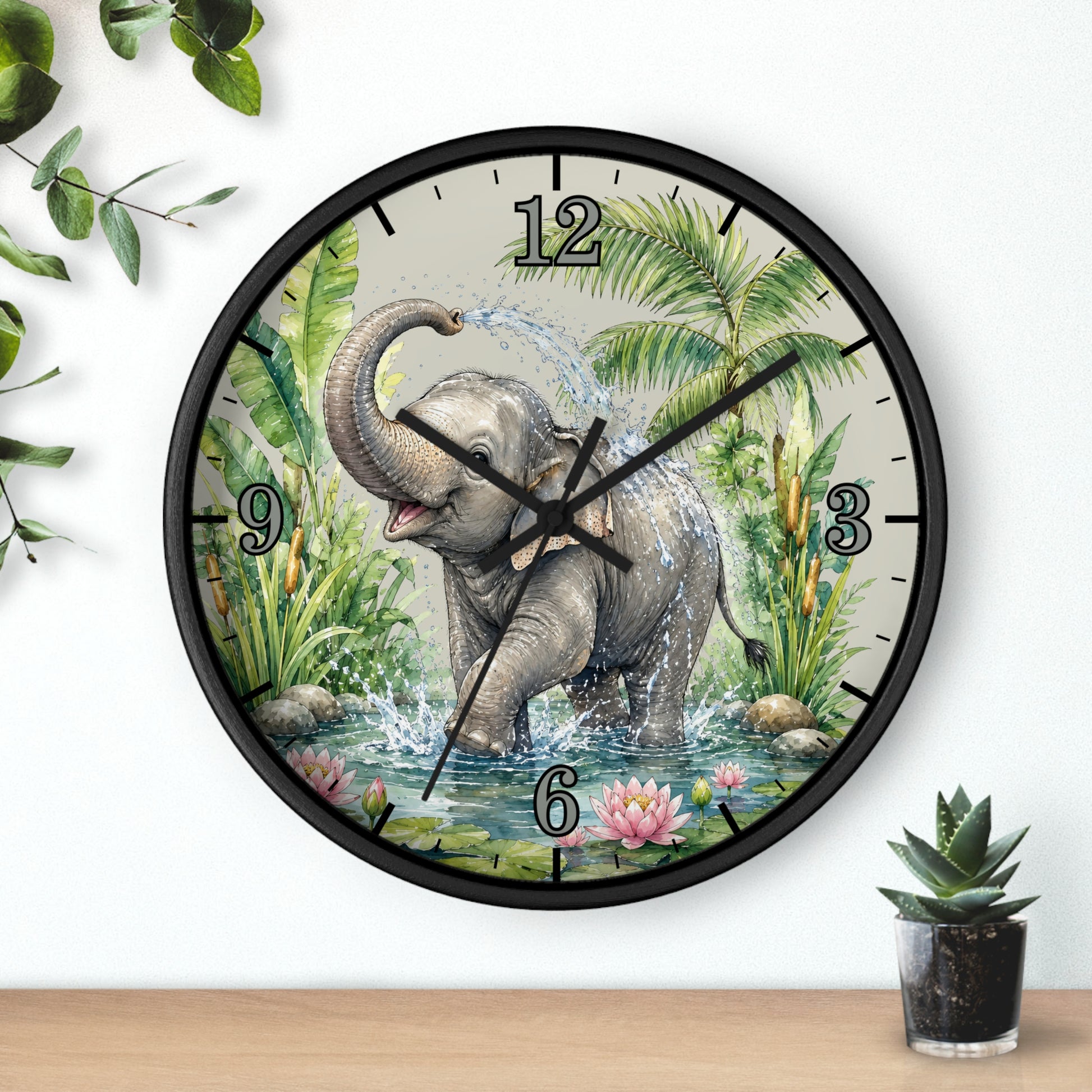 A round wall clock with a black frame and a printed illustration of an elephant bathing in water, surrounded by tropical plants. The clock is mounted on a white wall above a wooden surface with small potted plants nearby.