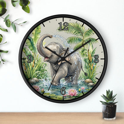 A round wall clock with a black frame and a printed illustration of an elephant bathing in water, surrounded by tropical plants. The clock is mounted on a white wall above a wooden surface with small potted plants nearby.