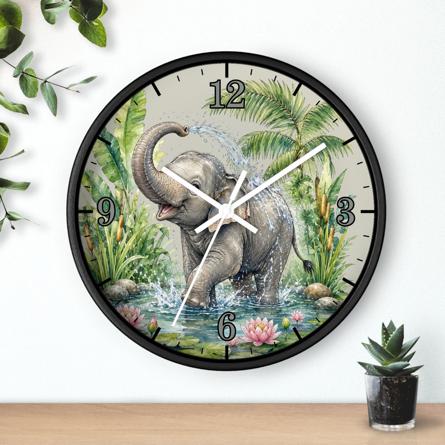 A round wall clock with a black frame and a printed illustration of a baby elephant standing in water with palm leaves and flowers. The clock is mounted on a white wall above a wooden surface, with decorative green leaves and a small potted succulent nearby.