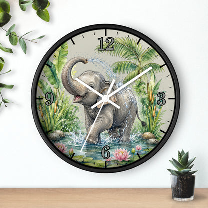 A round wall clock with a black frame and a printed illustration of a baby elephant standing in water with palm leaves and flowers. The clock is mounted on a white wall above a wooden surface, with decorative green leaves and a small potted succulent nearby.