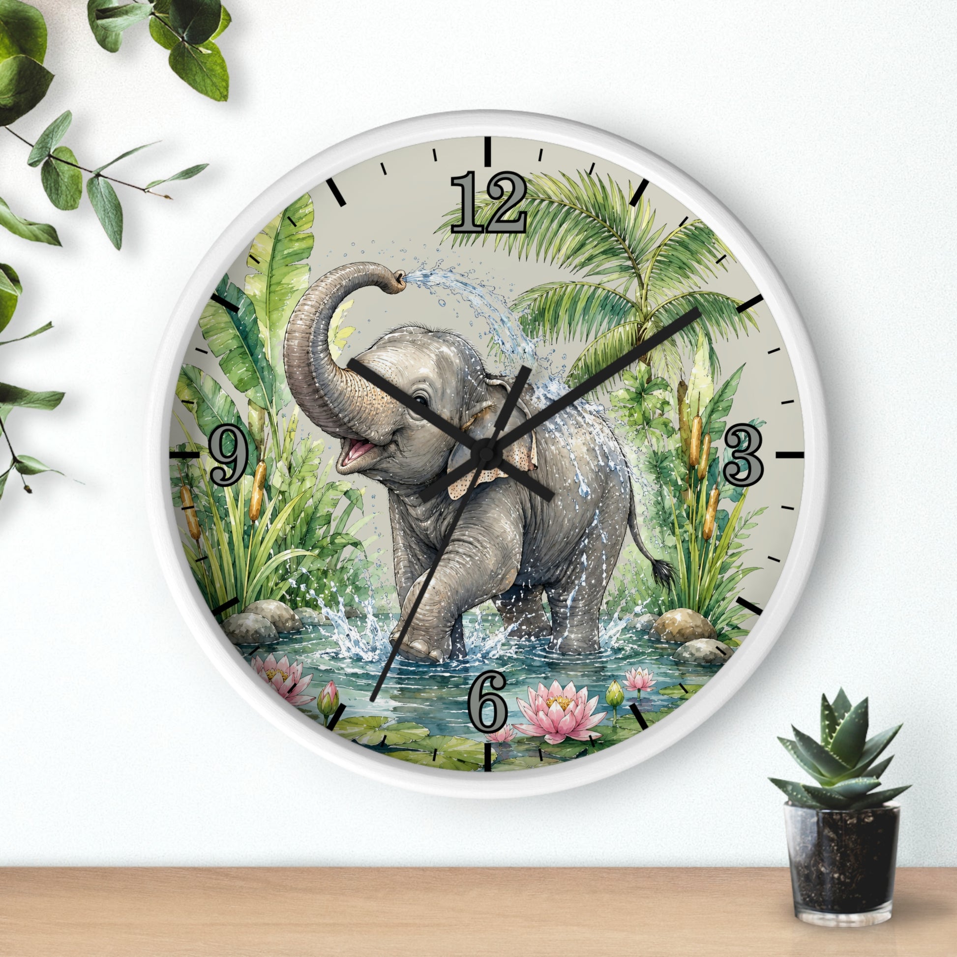 A round wall clock with a white frame and a printed illustration of a gray elephant standing in water among green tropical plants. The clock is mounted on a white wall above a light wood surface, with small green plants in the corners.