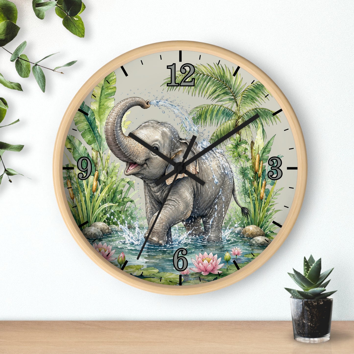 A round wall clock with a light wood frame and a printed illustration of a baby elephant standing in water with palm leaves and flowers. The clock is mounted on a white wall above a wooden surface, with decorative green leaves and a small potted succulent nearby.