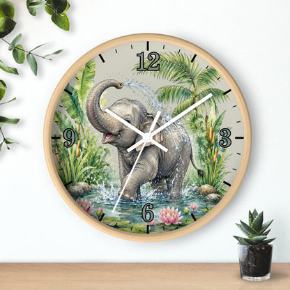 Round wall clock with a light wood frame and a printed illustration of an elephant standing in water among tropical plants. The clock is mounted on a white wall above a wooden surface, with a small potted succulent nearby and decorative leaves in the upper left corner.