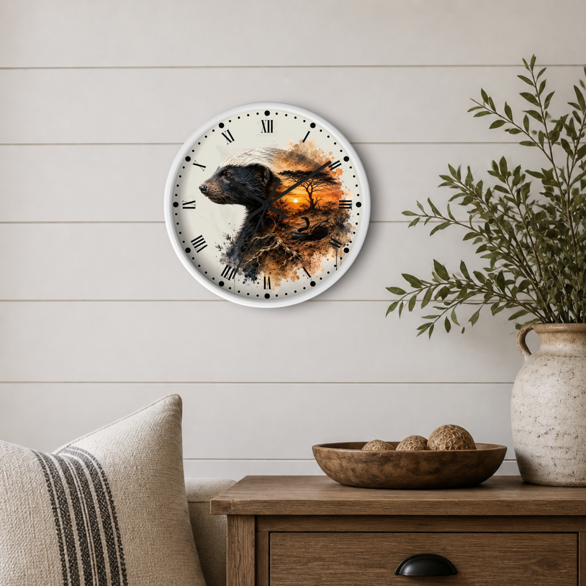 A round wall clock with a white frame and Roman numerals hangs on a light beige shiplap wall above a wooden side table. The clock face features a stylized badger silhouette blending into an orange and brown autumn landscape.