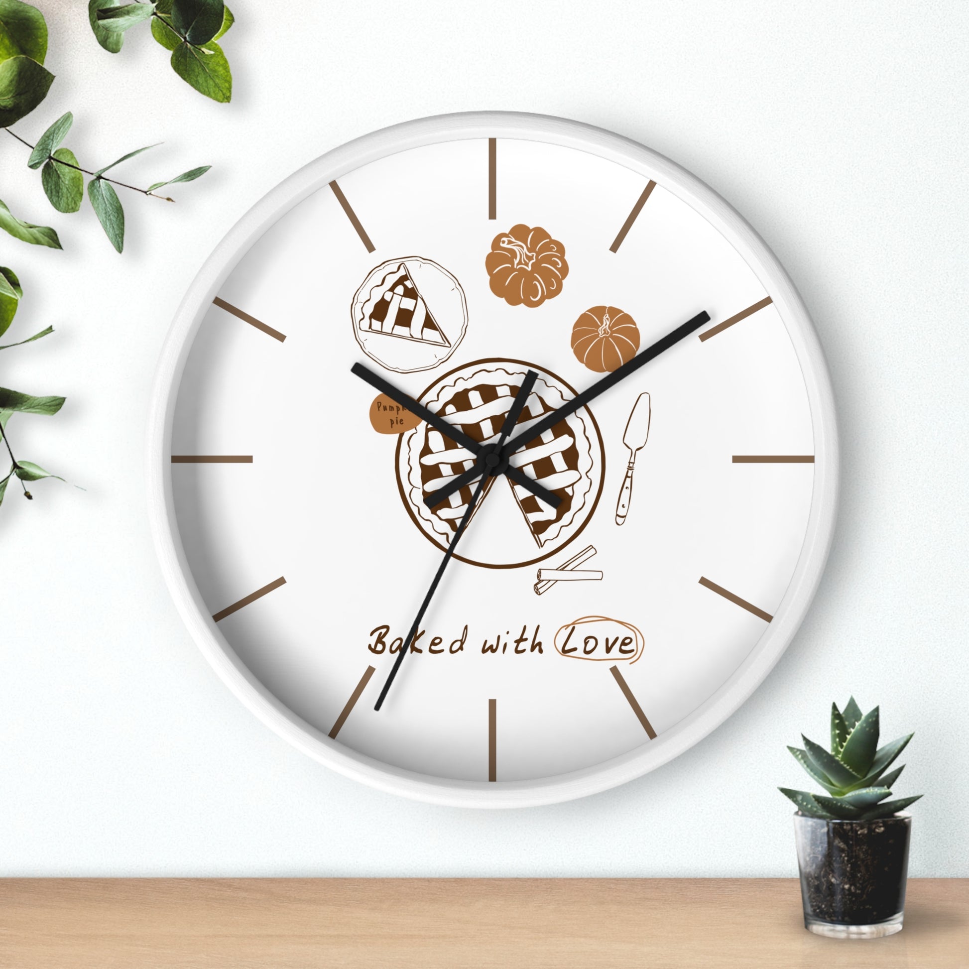 A round white wall clock with a baking-themed dial, featuring illustrations of a pie, pumpkin, cake slice, spatula, and the phrase 'Baked with Love' in brown. The clock is styled on a light wood surface against a white wall with green plants in the corners.