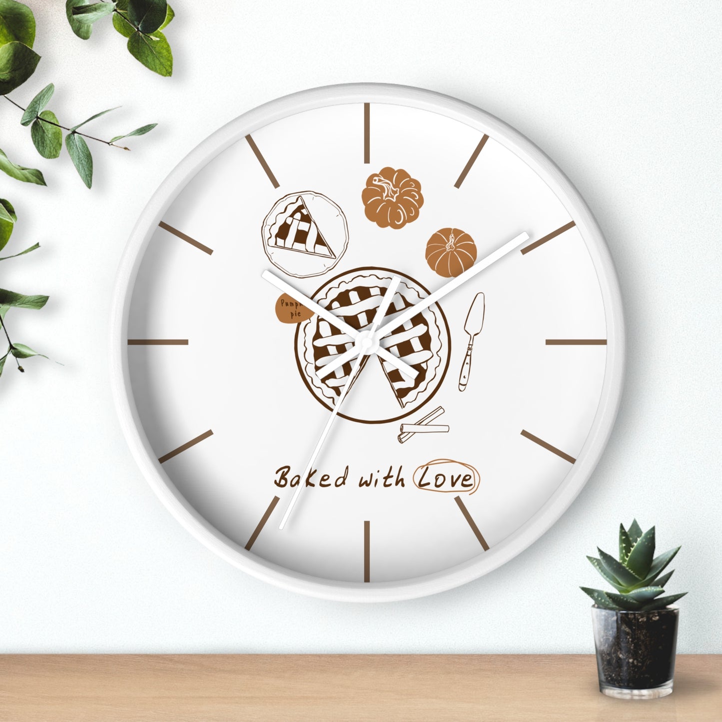 A round white wall clock with a baking-themed design, featuring illustrations of a pie, pumpkin, corn, and the text 'Baked with Love' in brown. The clock is styled on a light wood surface against a white wall with decorative green leaves and a small potted succulent.
