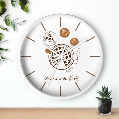 A round white wall clock with a baking-themed design, featuring illustrations of a pie, pumpkin, corn, and the text 'Baked with Love' in brown. The clock is styled on a light wood surface against a white wall with decorative green leaves and a small potted succulent.