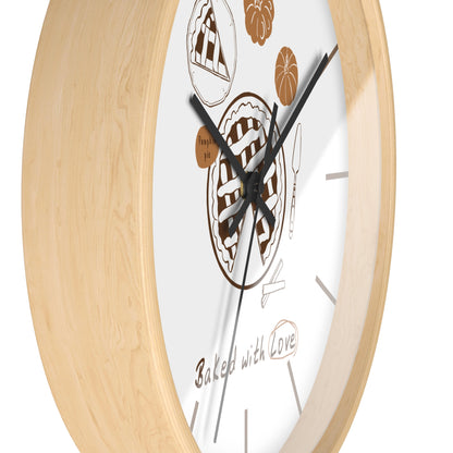 A close-up of a round wall clock with a light natural wood frame and a white face decorated with brown pie illustrations, black hands, and the phrase 'Baked with Love' near the bottom.