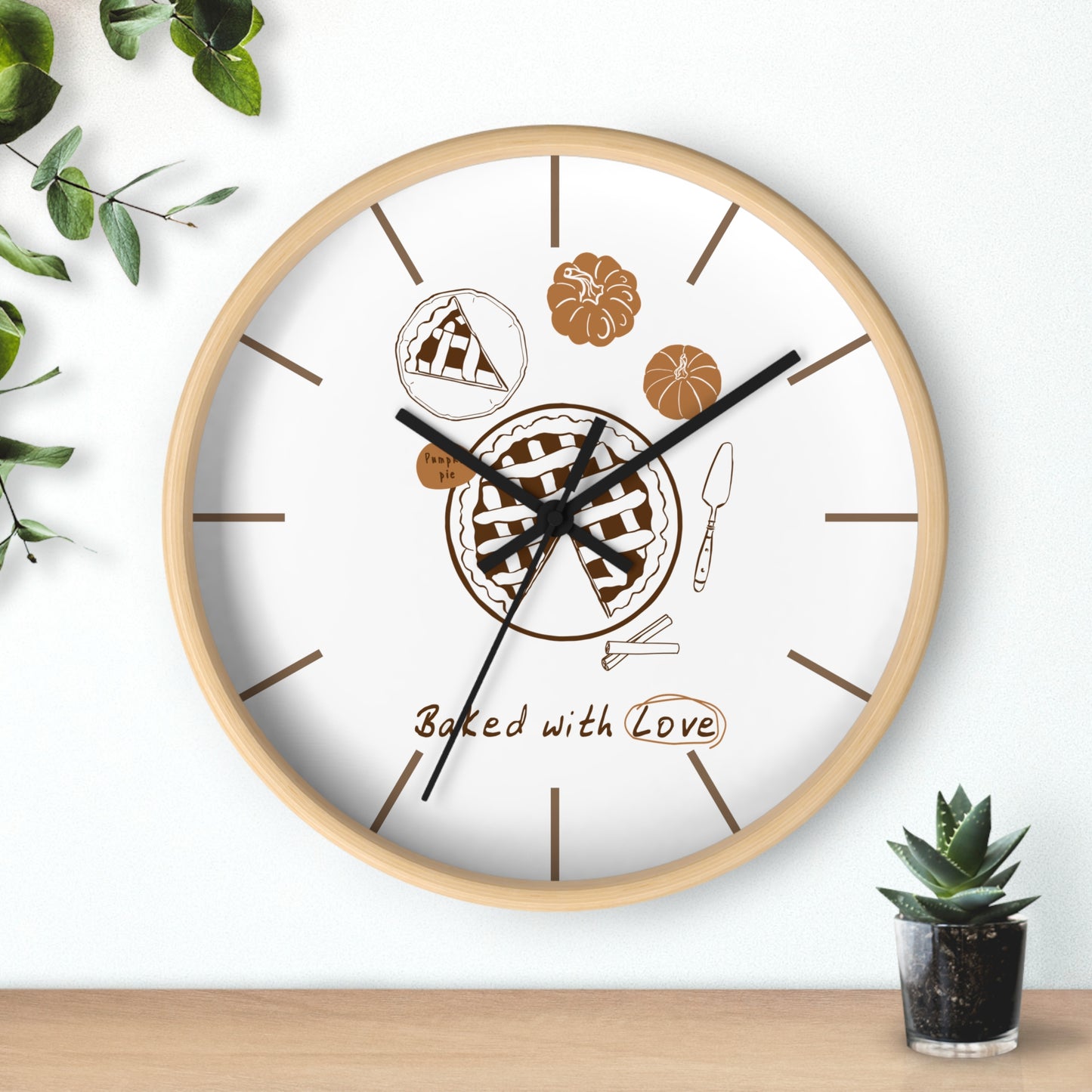 A round wall clock with a light wood frame and a white face, decorated with pie illustrations, brown hour markers, and the phrase 'Baked with Love' near the bottom. The clock is shown hanging on a white wall above a wooden surface, with small green plants in the corners.