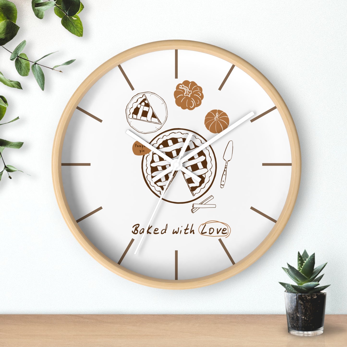 A round wall clock with a light wood frame and a white face, decorated with pie illustrations, brown line markers, and the words 'Baked with Love.' The clock is shown hanging on a white wall above a wooden surface, with small green plants in the corners.