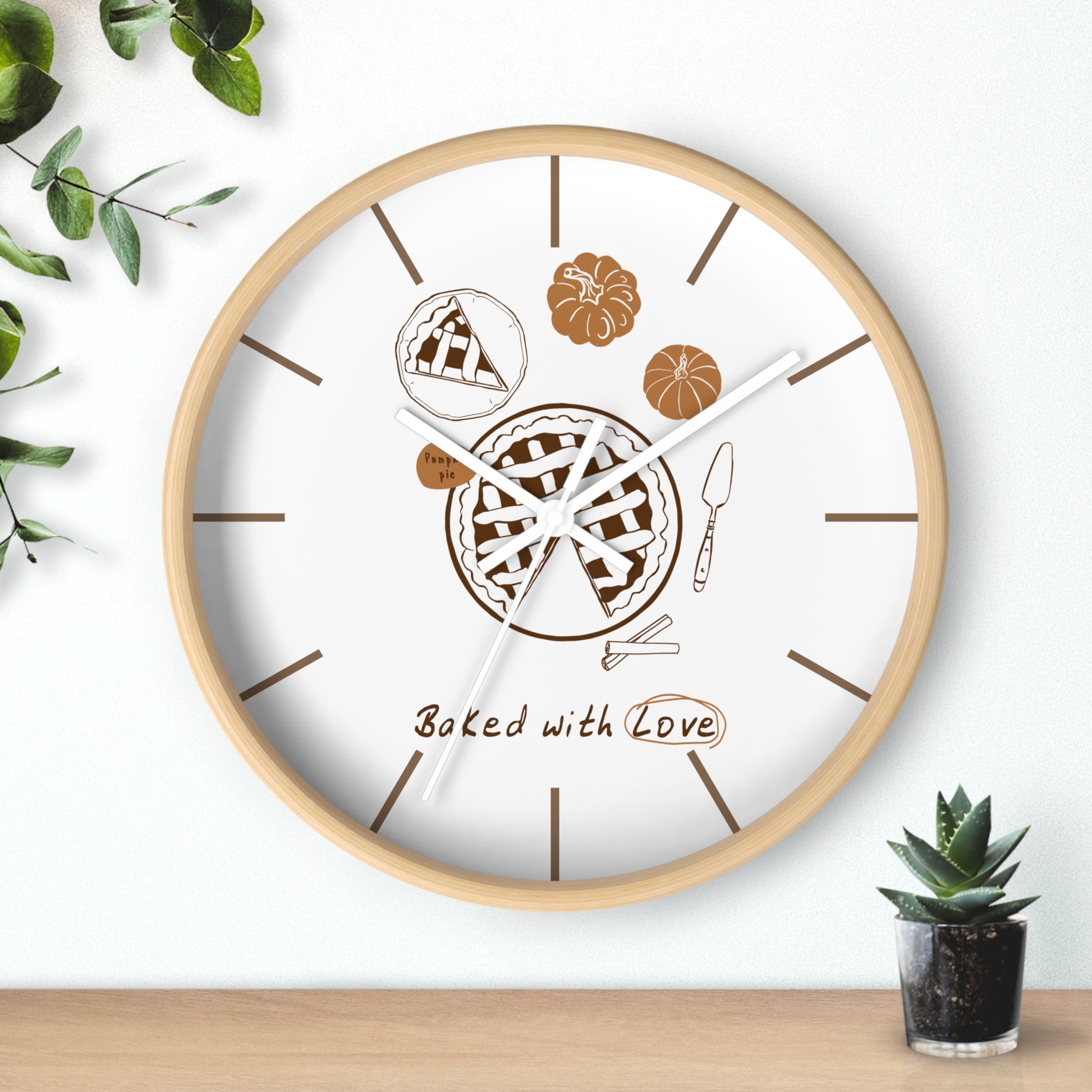 A round wall clock with a light wood frame and a white face, decorated with pie illustrations, brown line markers, and the words 'Baked with Love.' The clock is shown hanging on a white wall above a wooden surface, with small green plants in the corners.
