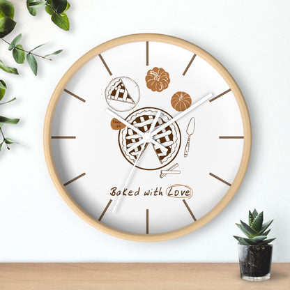 A round wall clock with a light wood frame and a white face, decorated with pie illustrations, brown line markers, and the words 'Baked with Love.' The clock is shown hanging on a white wall above a wooden surface, with small green plants in the corners.