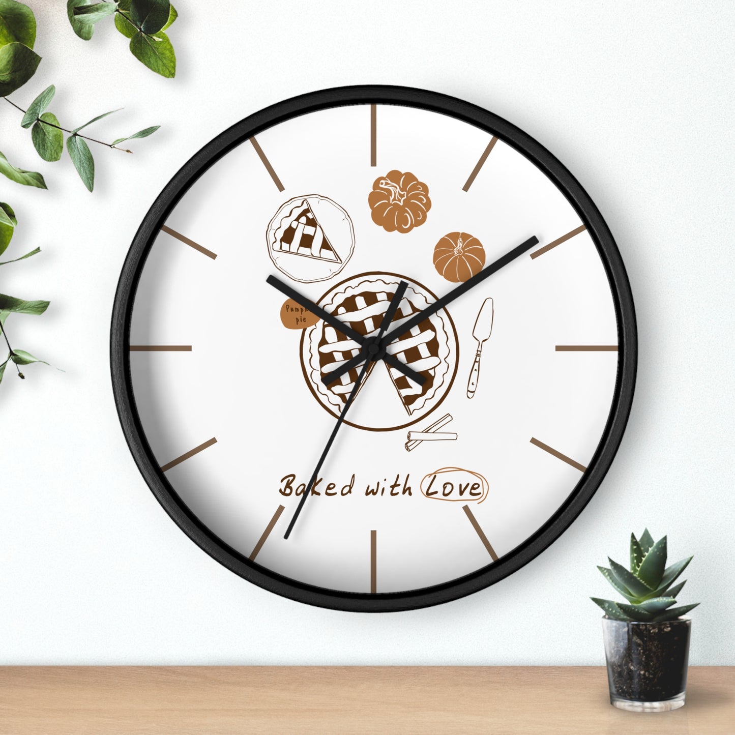A round wall clock with a black frame and white face, featuring baked pie illustrations and the words 'Baked with Love' on the dial. The clock is mounted on a white wall above a light wood surface, with small green plants in the corners.