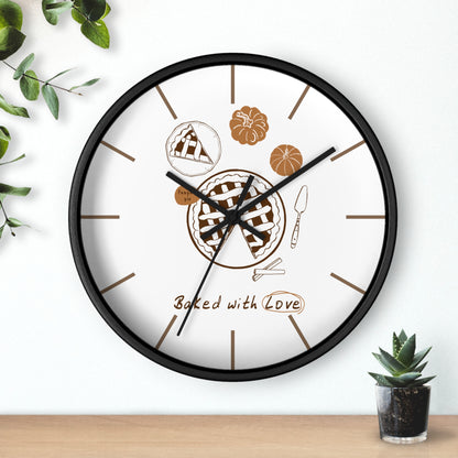 A round wall clock with a black frame and white face, featuring baked pie illustrations and the words 'Baked with Love' on the dial. The clock is mounted on a white wall above a light wood surface, with small green plants in the corners.