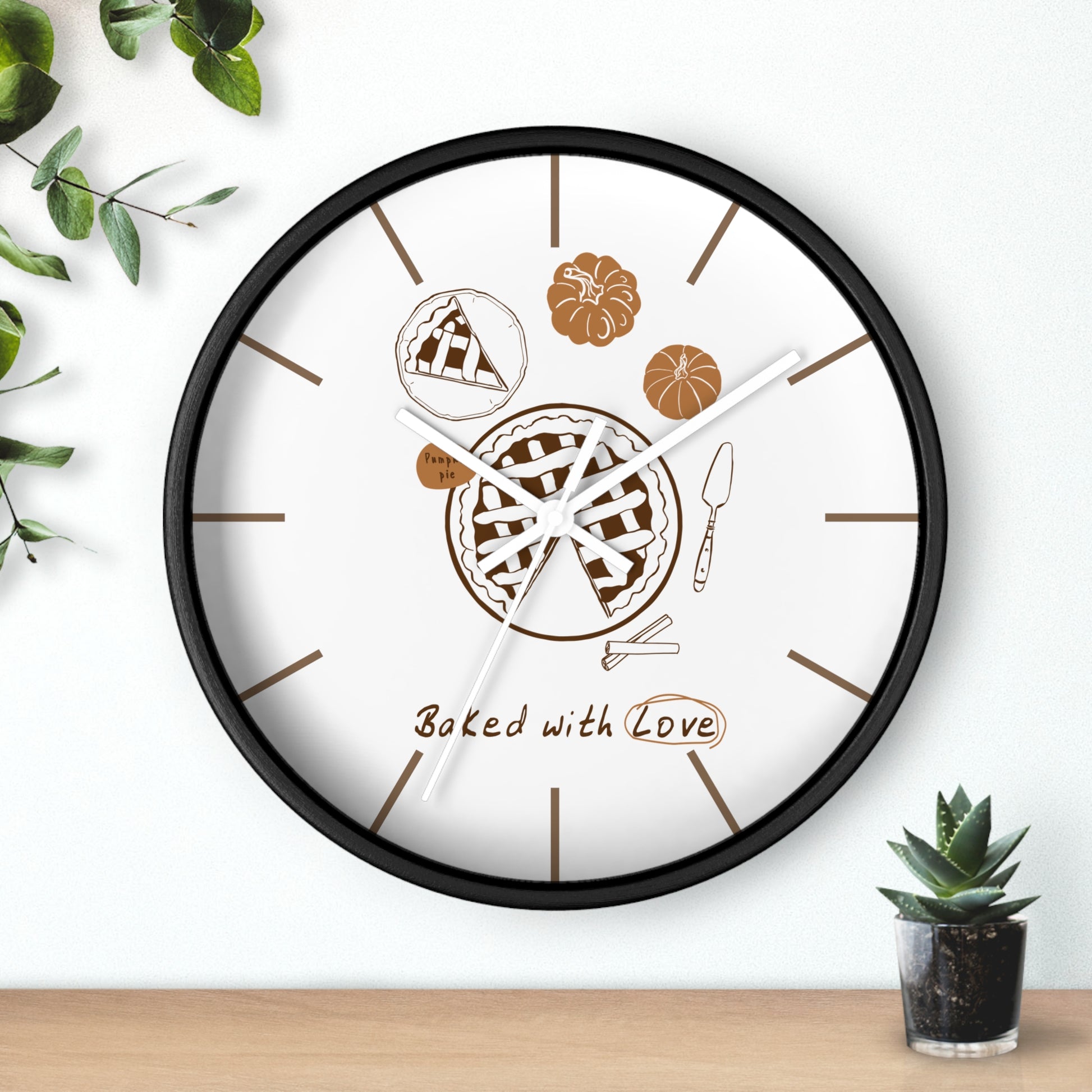 A round wall clock with a black frame and a white face, decorated with pie illustrations, brown line icons, and the text 'Baked with Love.' The clock is shown hanging on a white wall above a light wood surface, with small green plants in the corners.