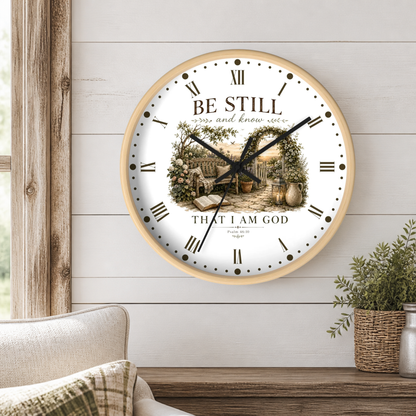 A round wall clock with a light wood frame hangs on a white shiplap wall above a wooden table. The clock face is white with black Roman numerals, black hands, and a central illustration of a garden scene.