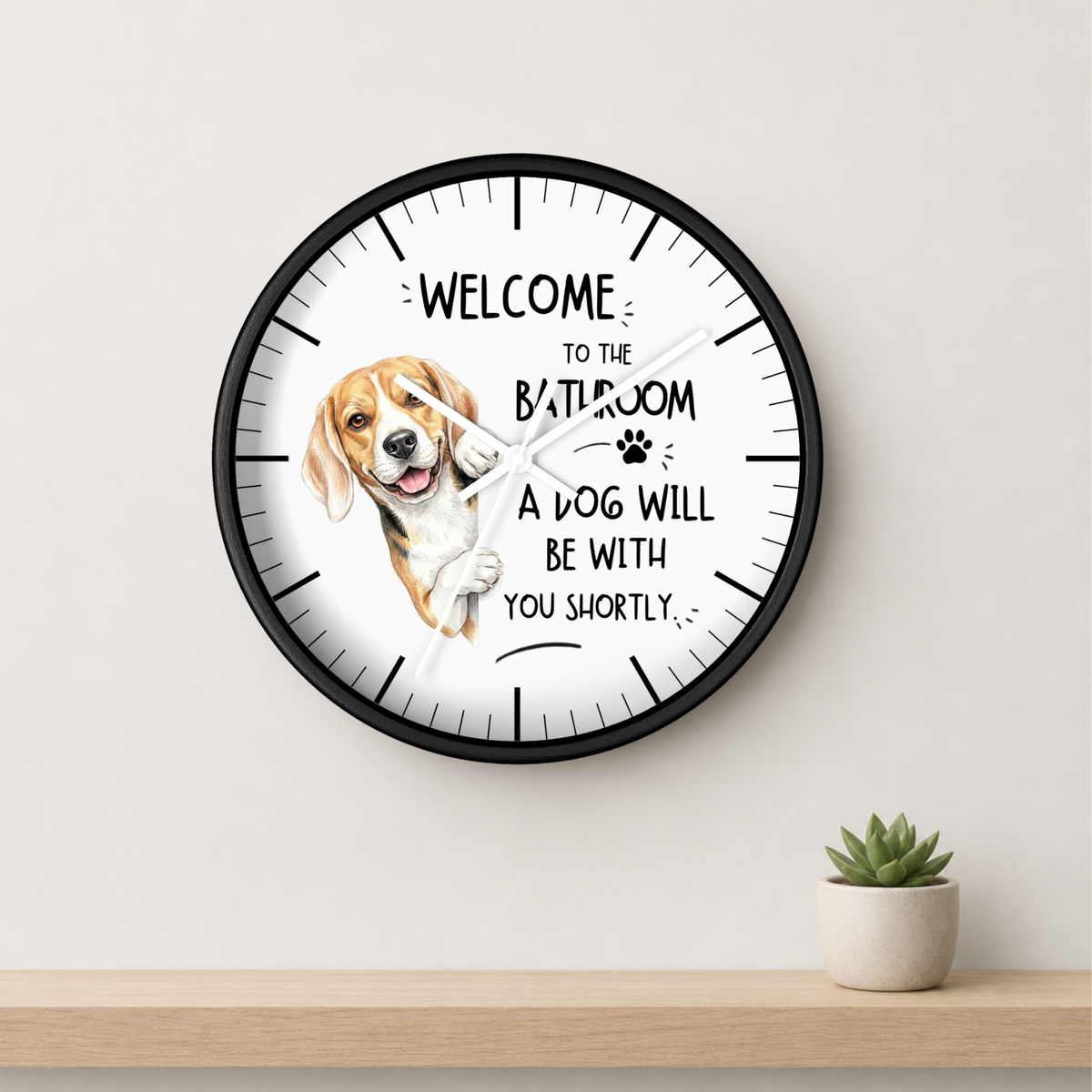 A round wall clock with a black frame and white face, featuring a beagle illustration and the text 'WELCOME TO THE BATHROOM A DOG WILL BE WITH YOU SHORTLY'. The clock is mounted on a light beige wall above a wooden shelf with a small potted succulent.