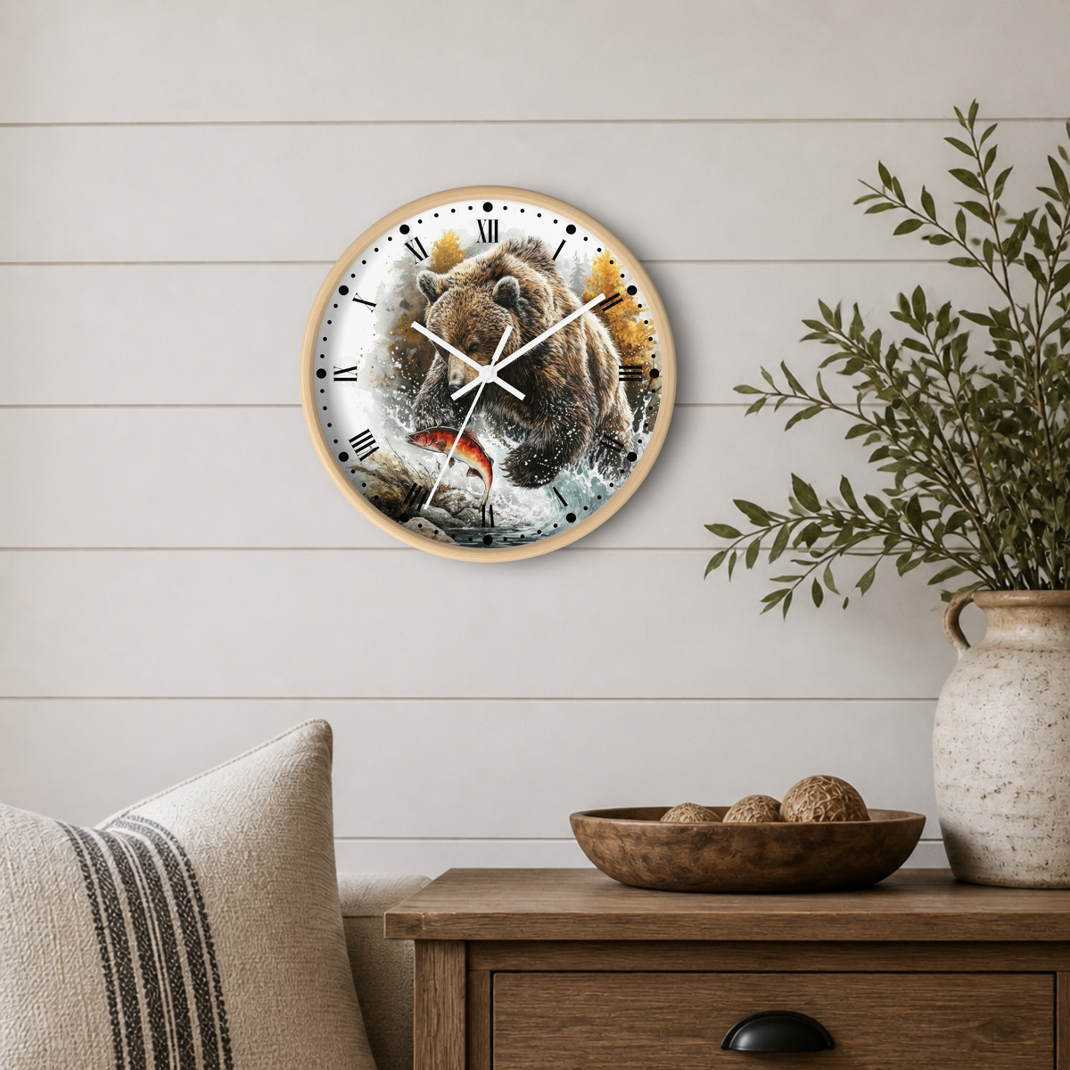 A round wall clock with a natural wood frame hangs on a white shiplap wall above a wooden side table. The clock face features a detailed watercolor-style illustration of a brown bear catching a red fish.