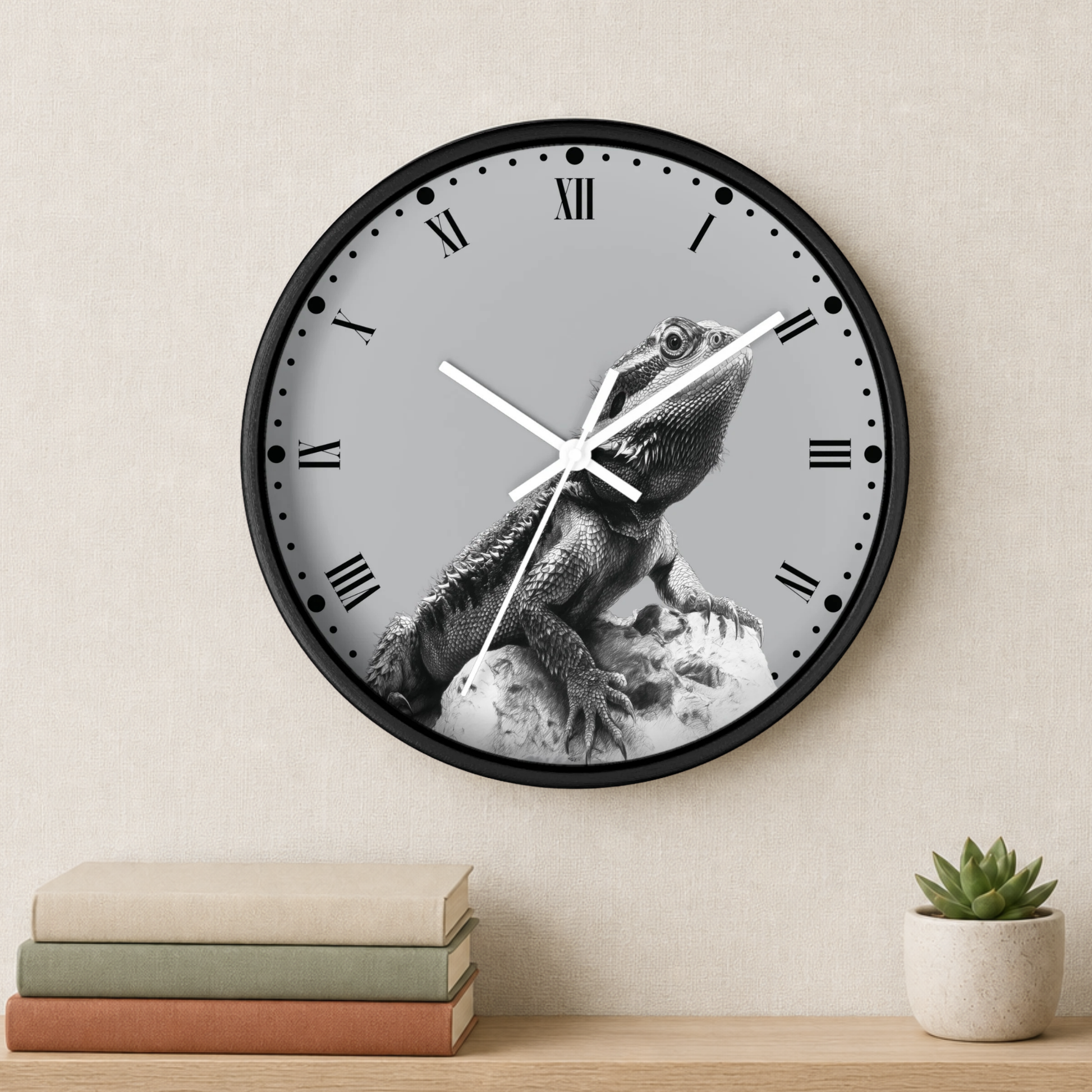 A round wall clock with a black frame and a gray dial featuring a black-and-white photo of a lizard on a rock. The clock is mounted on a beige wall above a wooden shelf with stacked books and a small potted succulent.