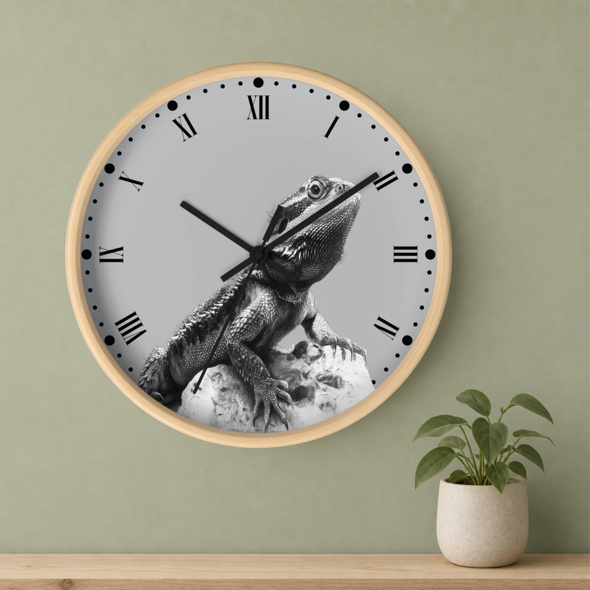 A round wall clock with a light wood frame and a gray face featuring a black-and-white bearded dragon illustration. The clock is mounted on a sage green wall above a wooden shelf, with a small potted green plant on the right.