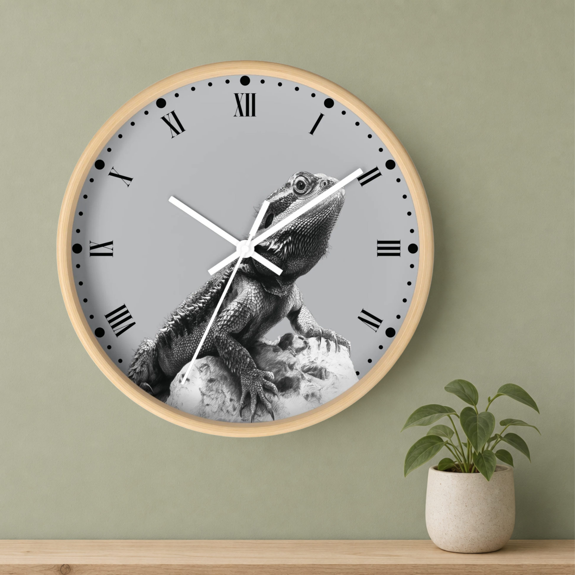A round wall clock with a light wood frame and a gray face featuring a black-and-white bearded dragon illustration. The clock is mounted on a sage green wall above a wooden shelf, with a small potted plant to the right.