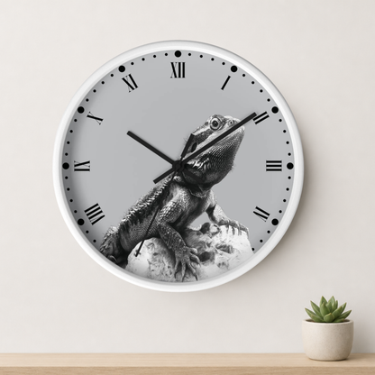 A round wall clock with a white frame and a grayscale lizard graphic on the face, mounted on a light wall above a small potted succulent.