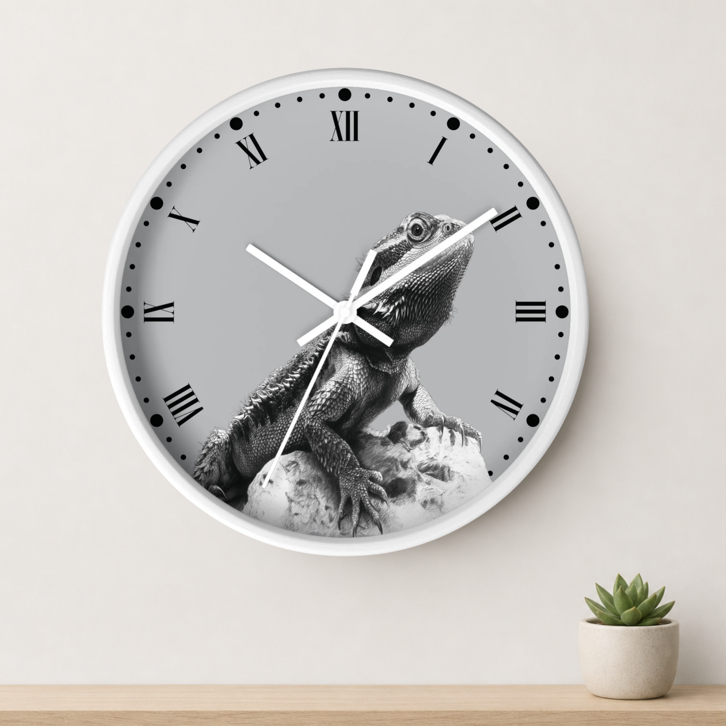 A round wall clock with a white frame and a gray dial featuring a black-and-white photo of a bearded dragon lizard. The clock is mounted on a light beige wall above a wooden surface with a small potted succulent.
