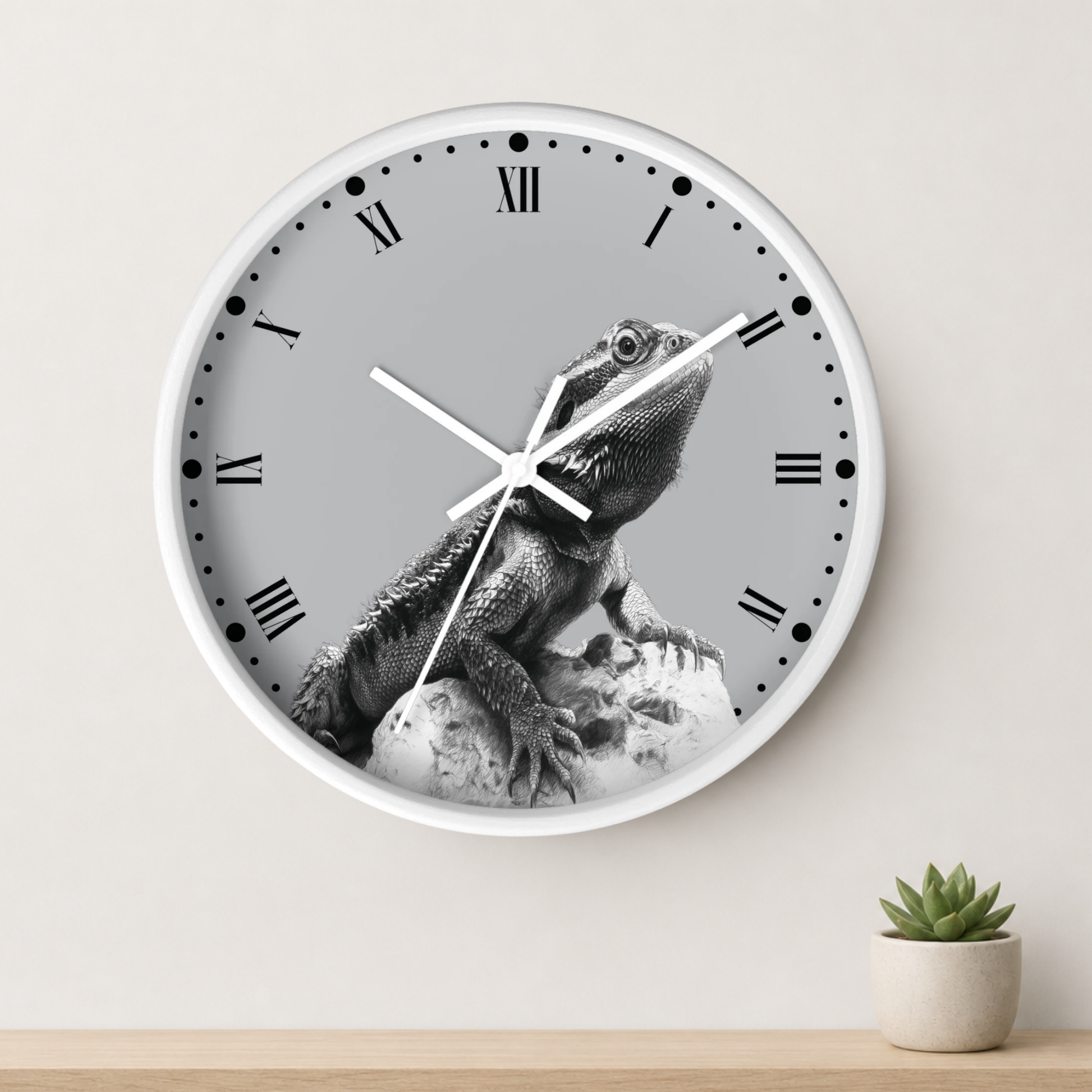 A round wall clock with a white frame and a gray dial featuring a black-and-white photo of a bearded dragon lizard. The clock is mounted on a light beige wall above a wooden surface with a small potted succulent.