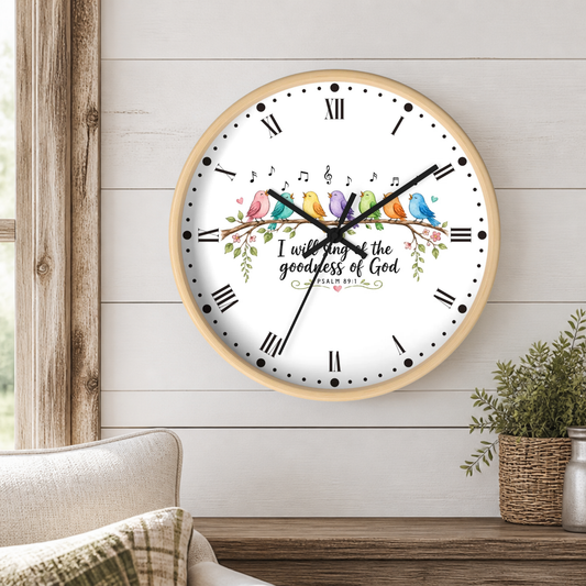 A round wall clock with a light wood frame and a white face, featuring four colorful birds, musical notes, floral details, and the text 'I wish every day of the goodness of God' with 'GLORY TO JESUS' below. The clock is mounted on a white shiplap wall above a wooden table with a small potted plant and part of a sofa visible.